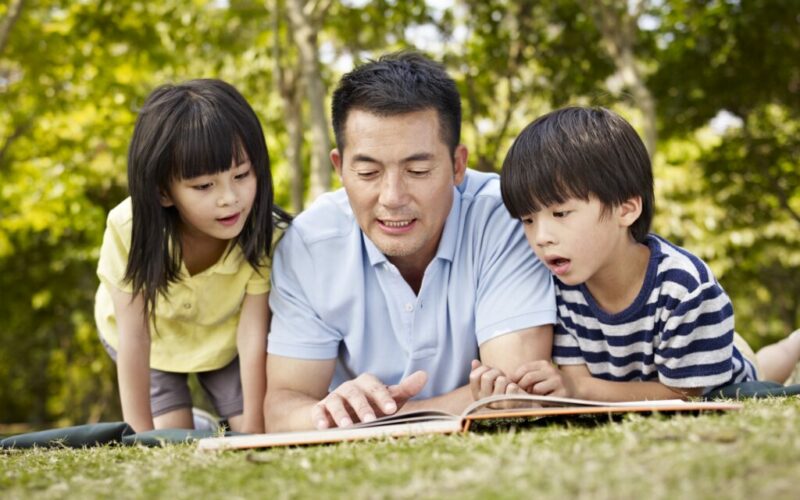 asian father and children lying on grass reading a book together in a park.